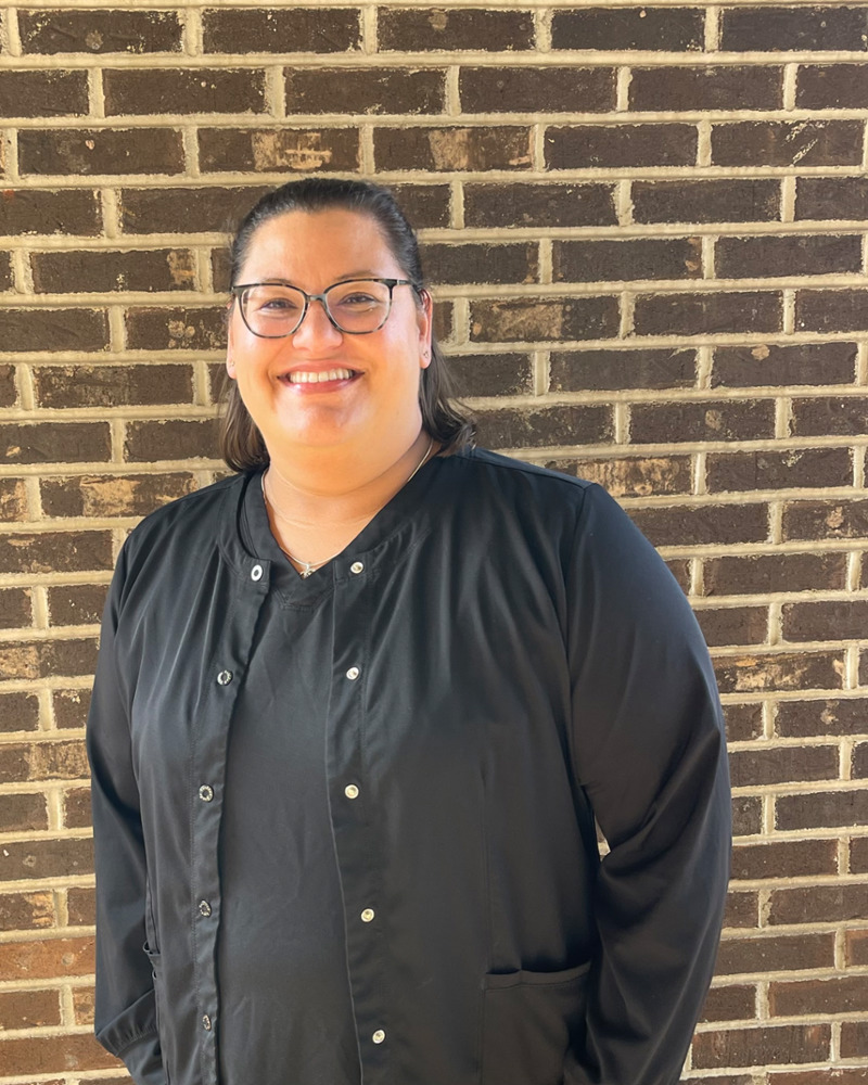 April, Insurance and Patient Care Coordinator, smiling warmly in black scrubs against a brick wall background