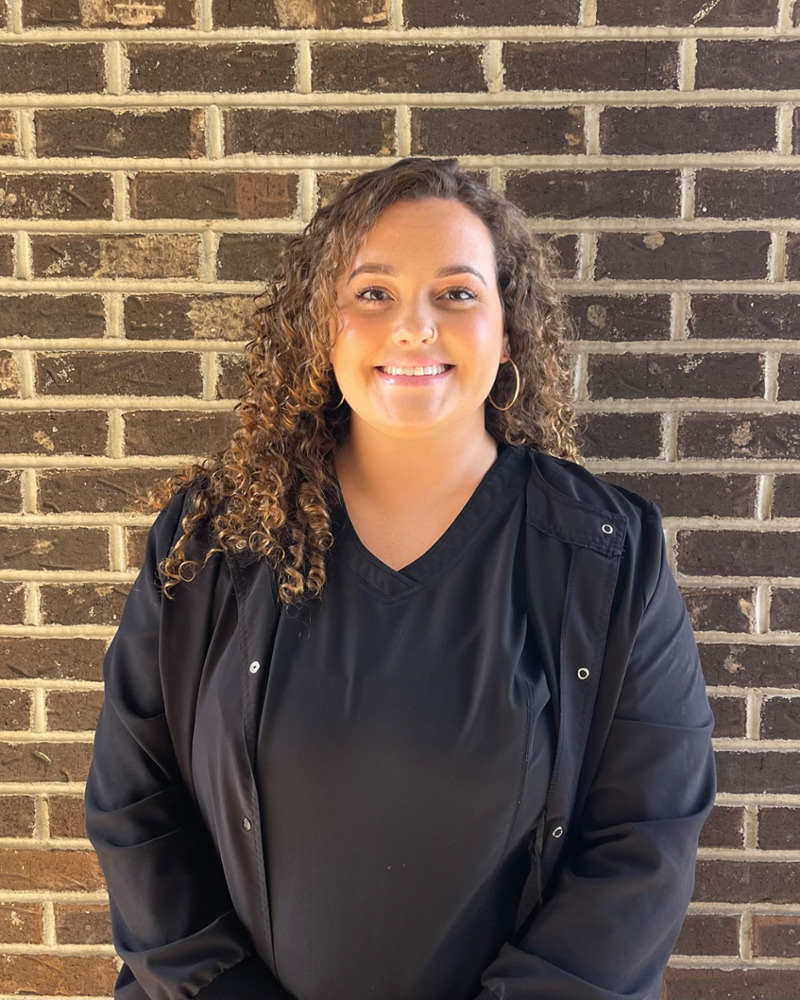 Destiny, registered dental assistant at Bryant Family Dentistry, smiling in black scrubs against a brick wall background